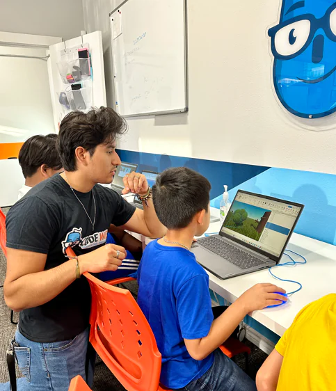 A Code Wiz instructor assists a young student with game development on a laptop in a classroom.