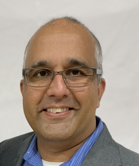 A smiling man with glasses, wearing a blue striped shirt and grey blazer.