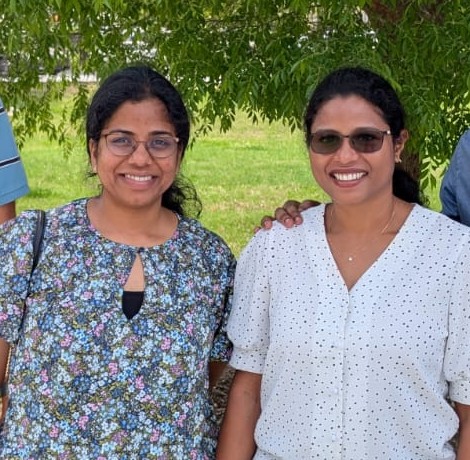 Two smiling women stand together outdoors with green trees in the background.