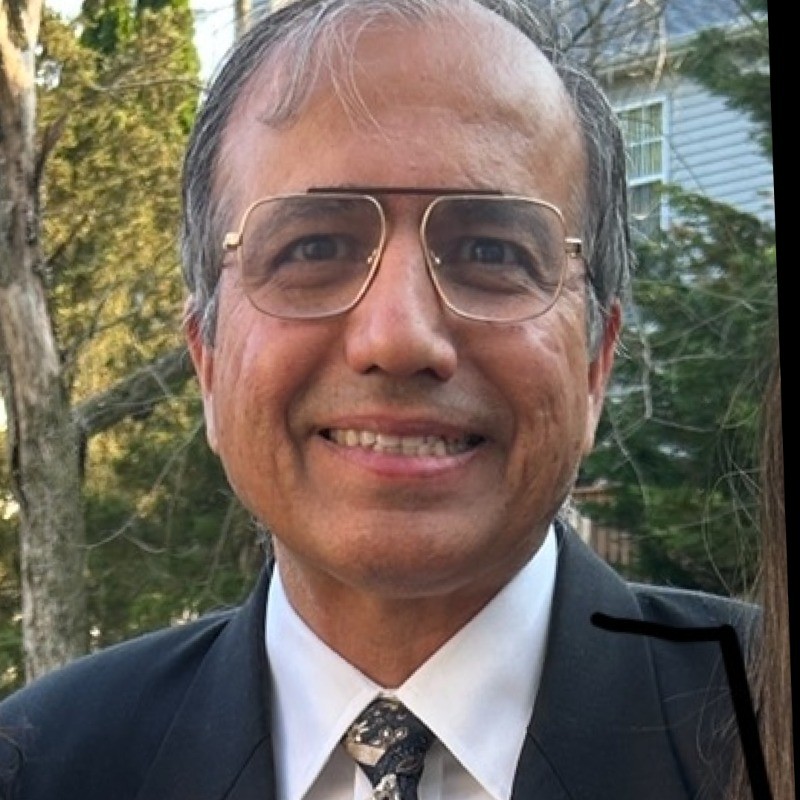Smiling man in a suit, tie, and glasses with a natural outdoor background. TITLE:
