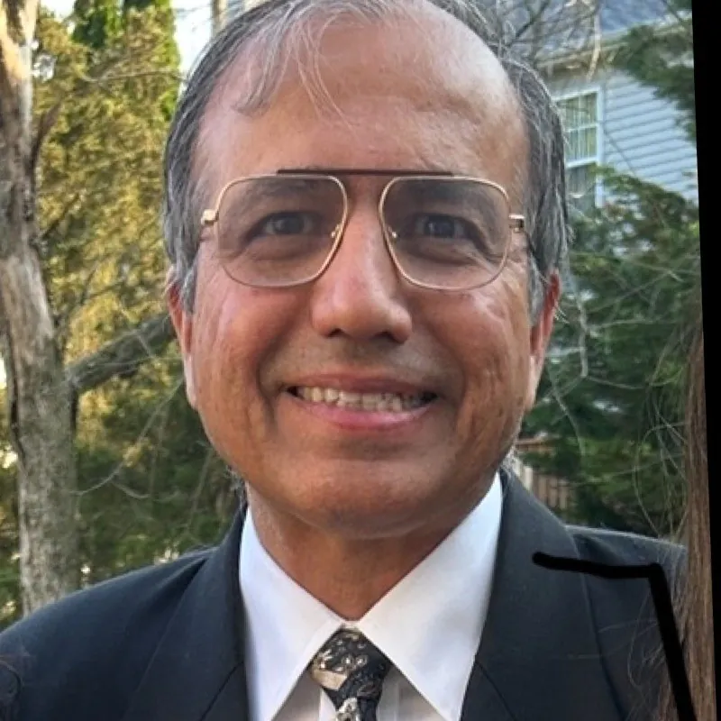 Smiling man in a suit, tie, and glasses with a natural outdoor background. TITLE: