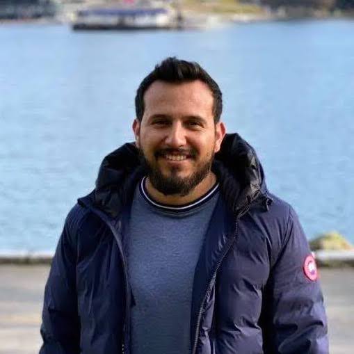 A smiling man with a beard wears a blue puffer jacket by the water. TITLE