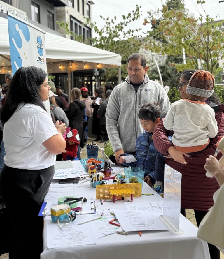 A Code Wiz representative talks to a family with children at an interactive STEM activity booth. TITLE