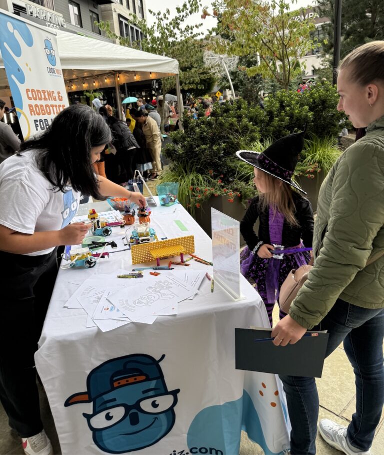 A Code Wiz instructor helps a young witch with coding and robotics activities at an outdoor fair.