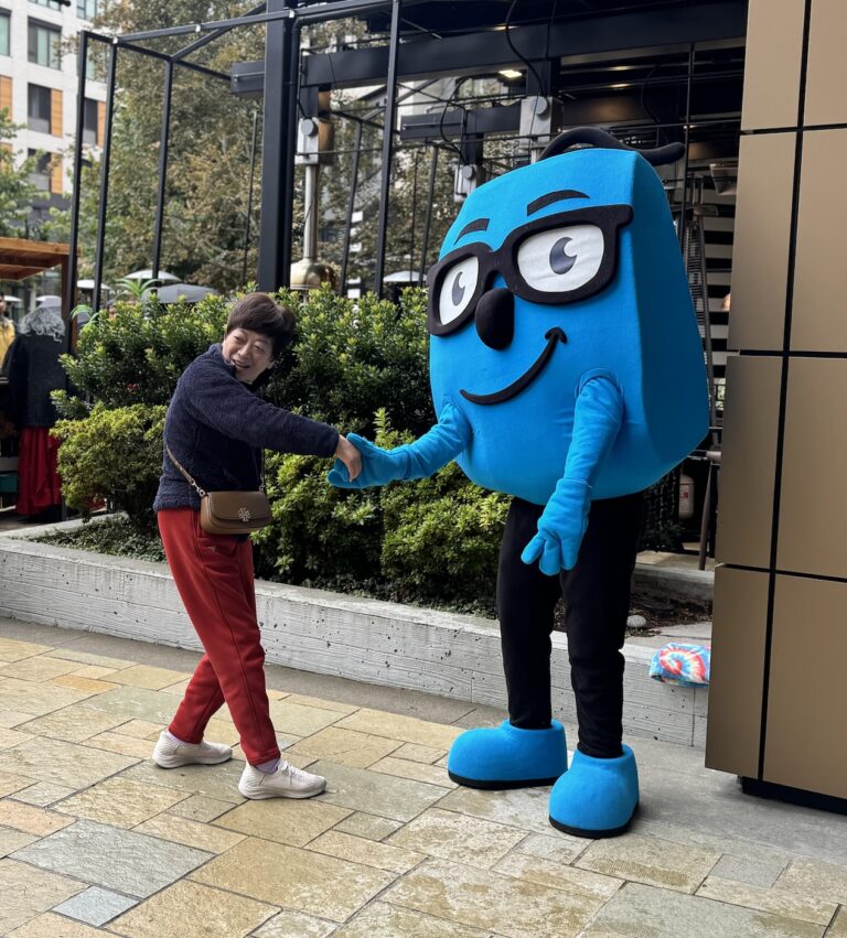 A happy woman shakes hands with a friendly blue mascot wearing glasses outdoors.