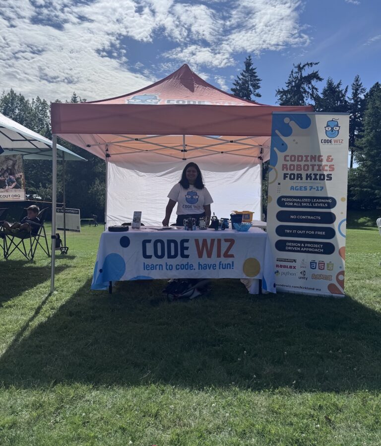 A smiling woman stands at a Code Wiz booth promoting coding and robotics for kids at an outdoor event