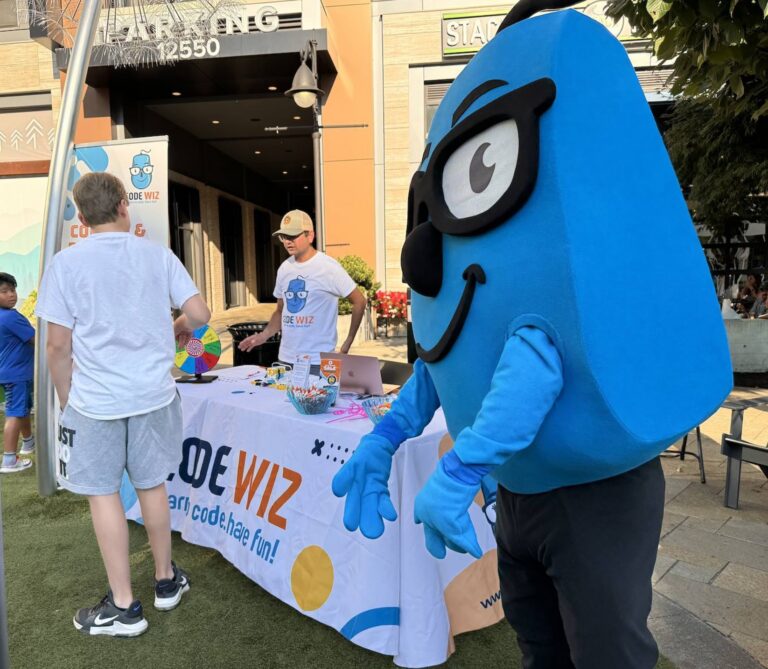 A friendly blue Code Wiz mascot stands next to an event table with a man and child.