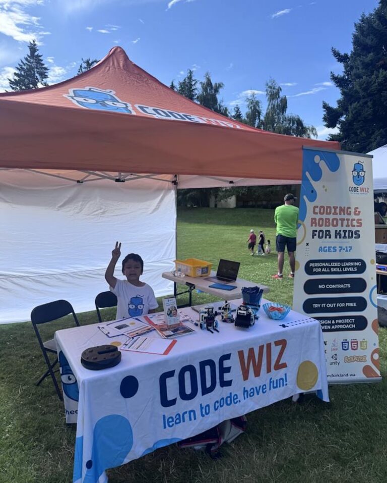 Smiling boy makes peace sign at a Code Wiz coding and robotics outdoor booth. TITLE: