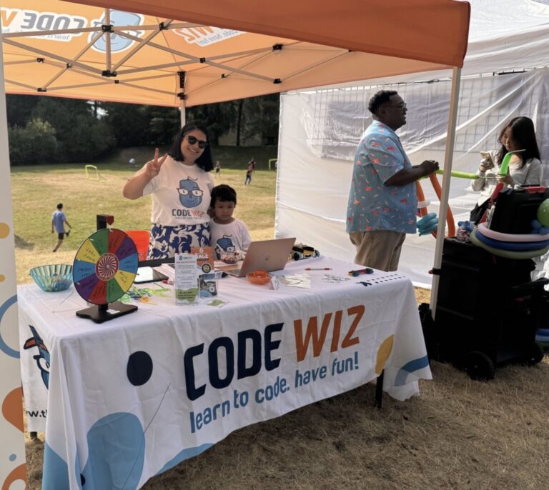 A smiling woman and child staff a Code Wiz booth with a spinning wheel and a balloon artist