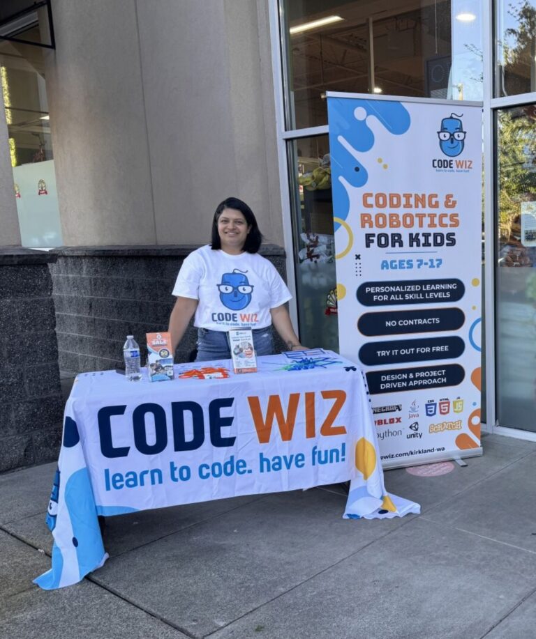 A smiling woman promotes Code Wiz coding and robotics for kids at an outdoor booth. TITLE: