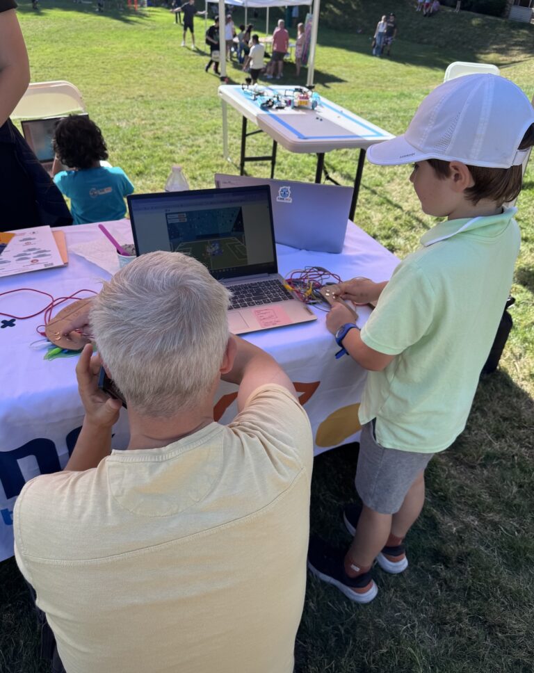A child and adult engage with a laptop and electronics at an outdoor educational event. TITLE
