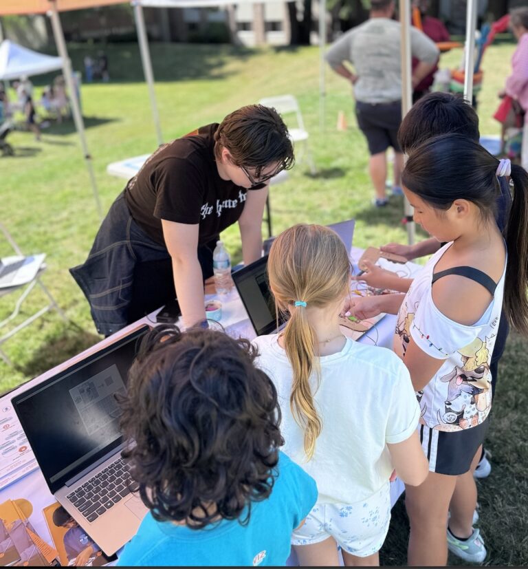 An adult helps children with a hands-on STEM project involving laptops and electronics at an outdoor event