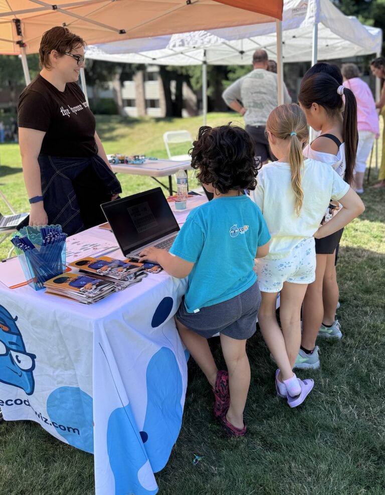 An instructor guides children using a laptop at an outdoor coding education booth.