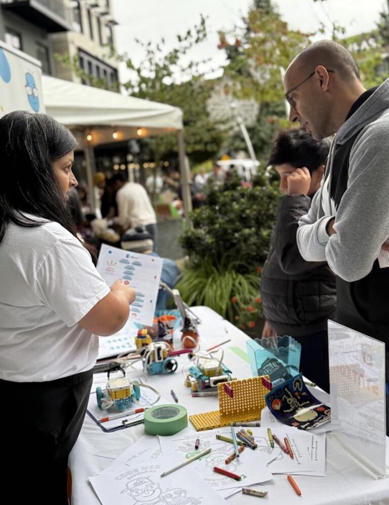 A woman explains robotics activities at an outdoor STEM education booth to a man and child