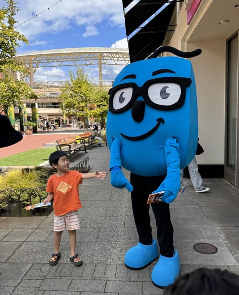 A child in an orange shirt reaches out to a friendly blue mascot in an outdoor setting.