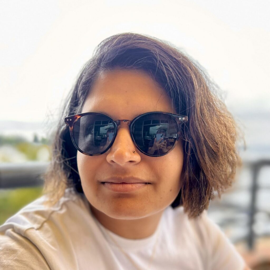 A person with short brown hair and a nose piercing wears sunglasses, reflecting a phone. TITLE