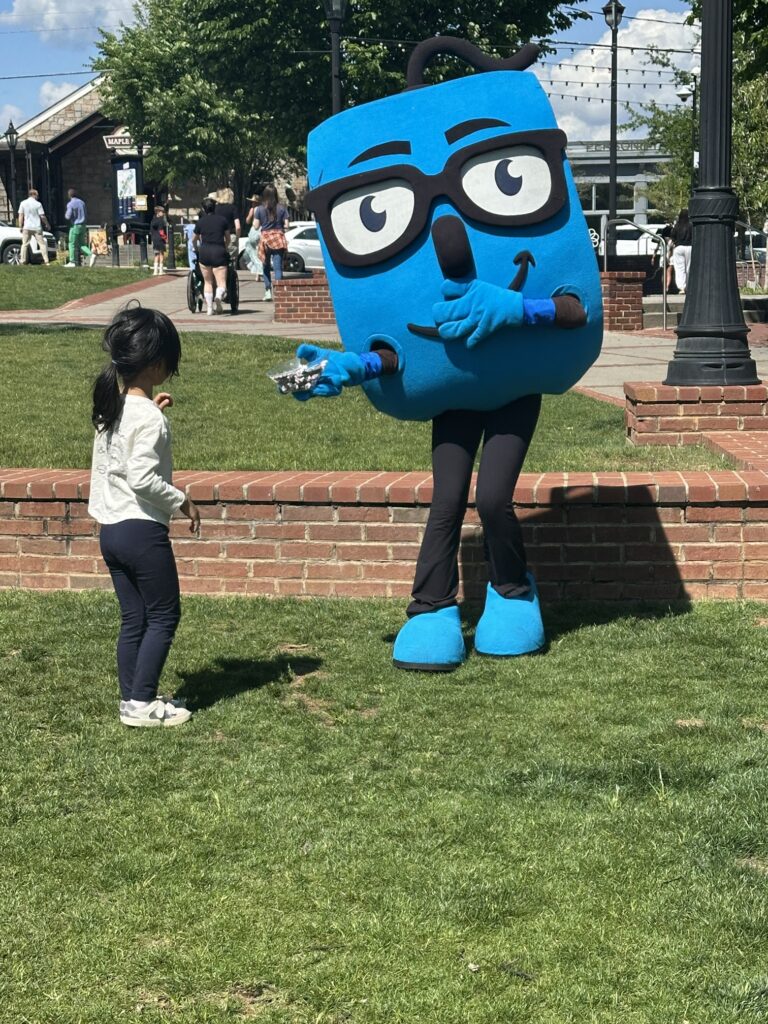 A friendly blue mascot with glasses offers a small treat to a young child on a sunny lawn.
