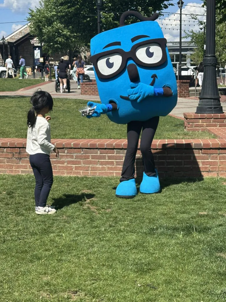 A friendly blue mascot with glasses offers a small treat to a young child on a sunny lawn.