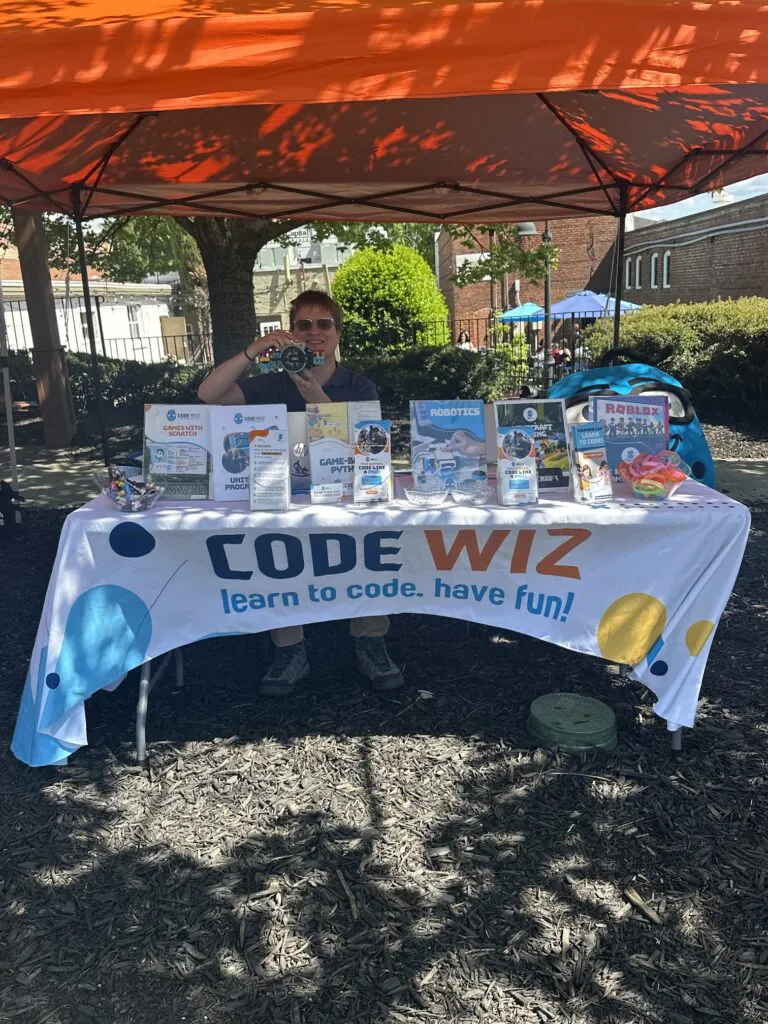 A person smiles behind a Code Wiz booth, displaying coding and robotics program information.