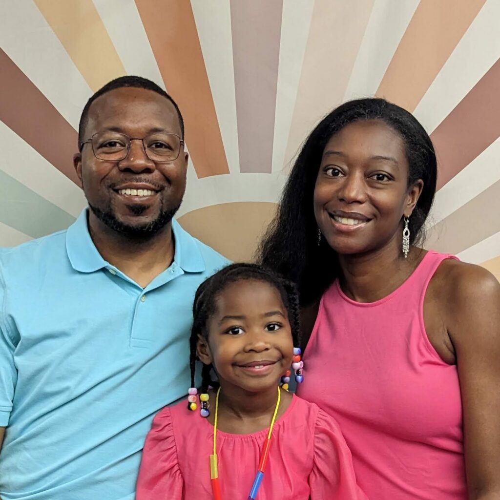 A smiling African American family of three stands together against a pastel striped background.