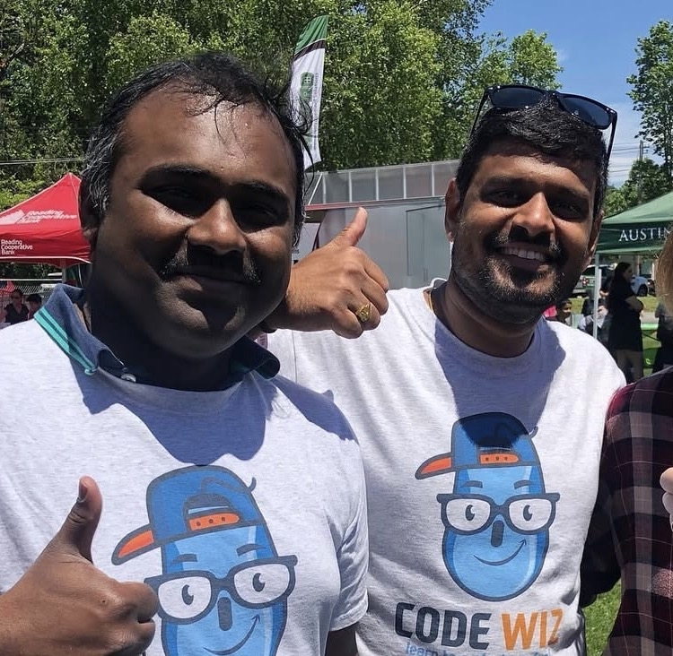 Two smiling men in Code Wiz t-shirts give thumbs up at an outdoor event.