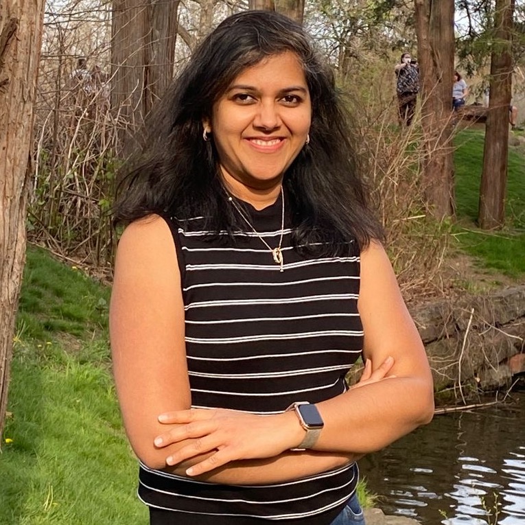 A smiling woman in a striped top and smartwatch stands with crossed arms in a sunny park.
