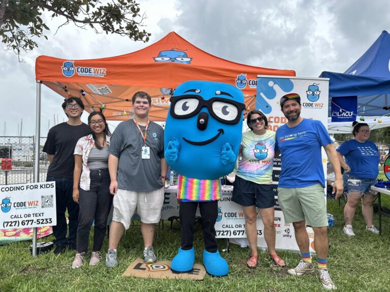 A cheerful Code Wiz team and their blue mascot pose outdoors under an orange tent. TITLE