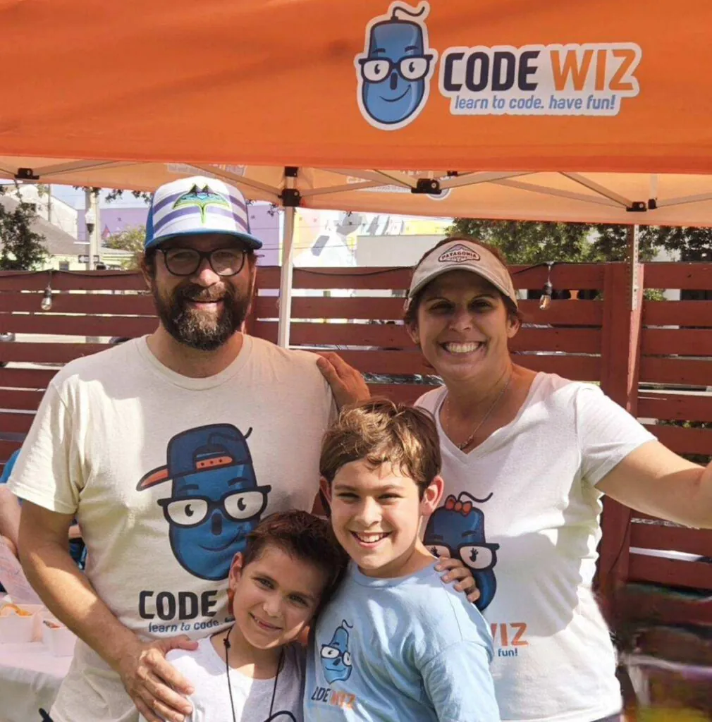A smiling family of four stands under an orange Code Wiz tent, promoting coding education.