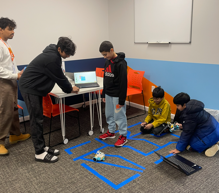 Kids and instructor learn robotics, programming small robots on a taped floor path.