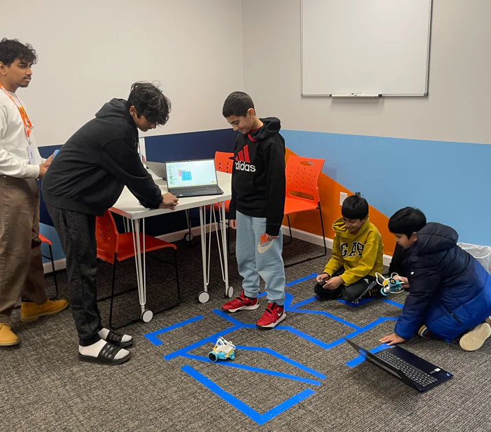 Kids and instructor learn robotics, programming small robots on a taped floor path.