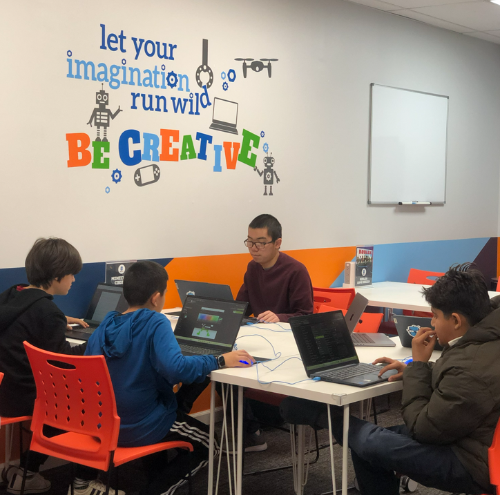 Students and an instructor code on laptops in a vibrant classroom with a "Be Creative