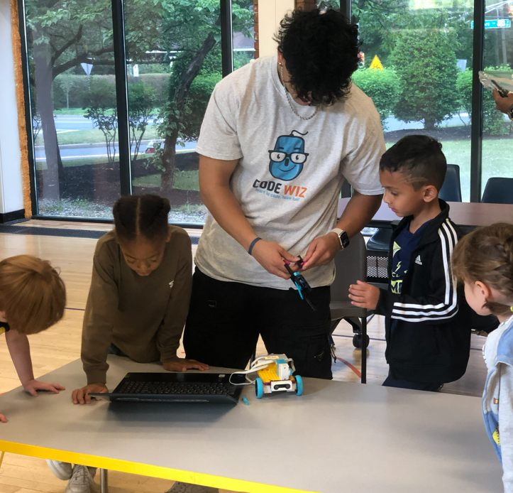 An instructor wearing a Code Wiz shirt teaches children coding and robotics with a laptop and robot.