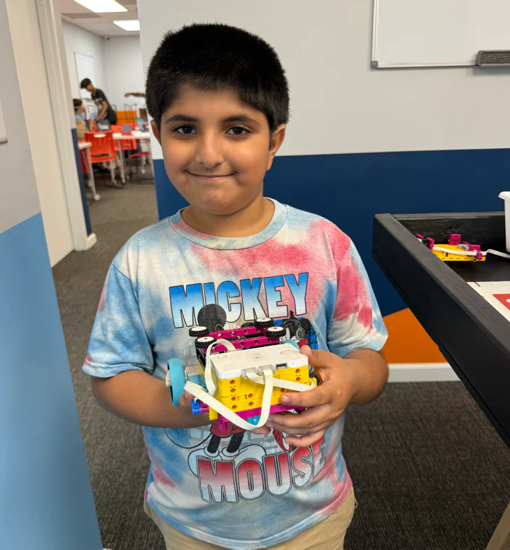A young boy proudly displays his colorful Lego robot in a STEM classroom.