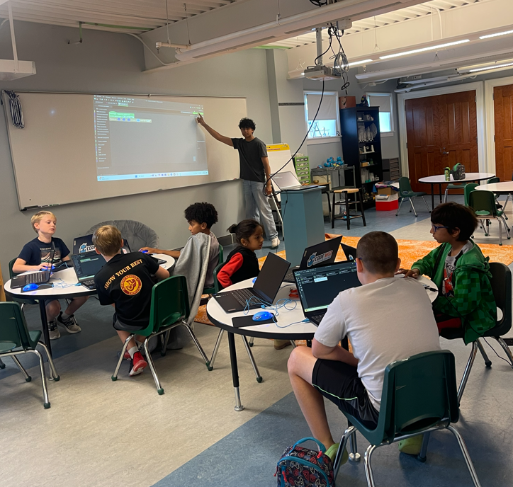 Young students code on laptops while an instructor teaches from a projected screen.
