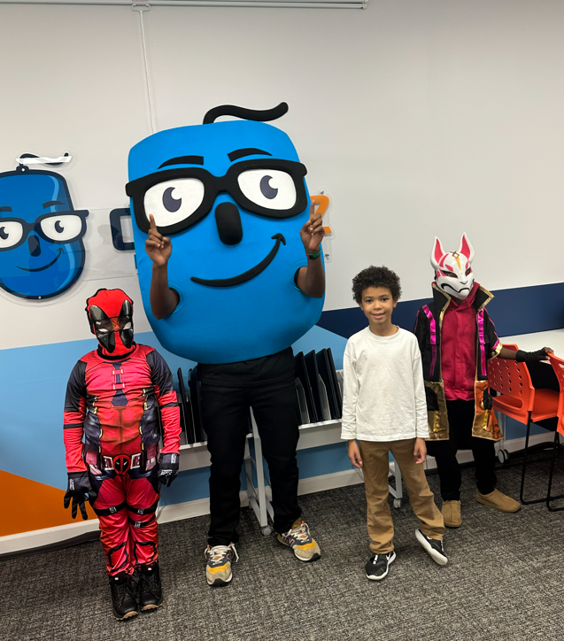 Three children in costumes pose with a smiling blue mascot character.