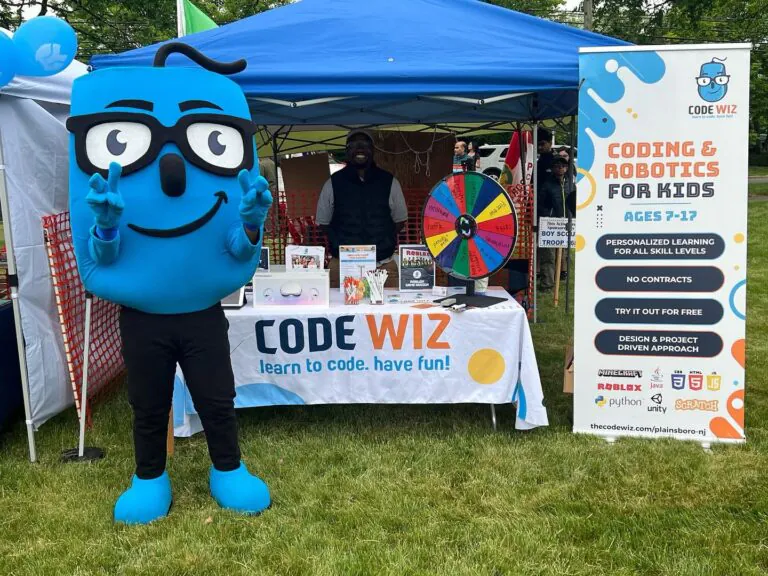 A smiling blue Code Wiz mascot poses at an outdoor booth for coding and robotics. TITLE
