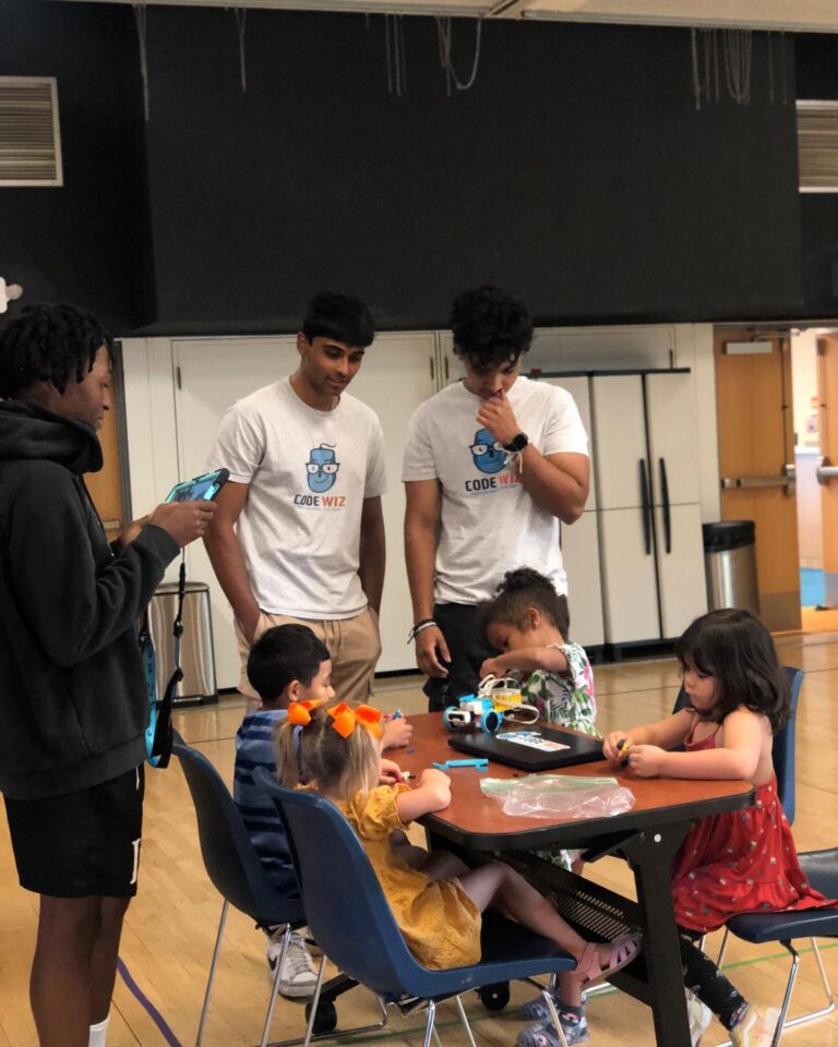 Young men in Code Wiz shirts teach children robotics at a table. TITLE: