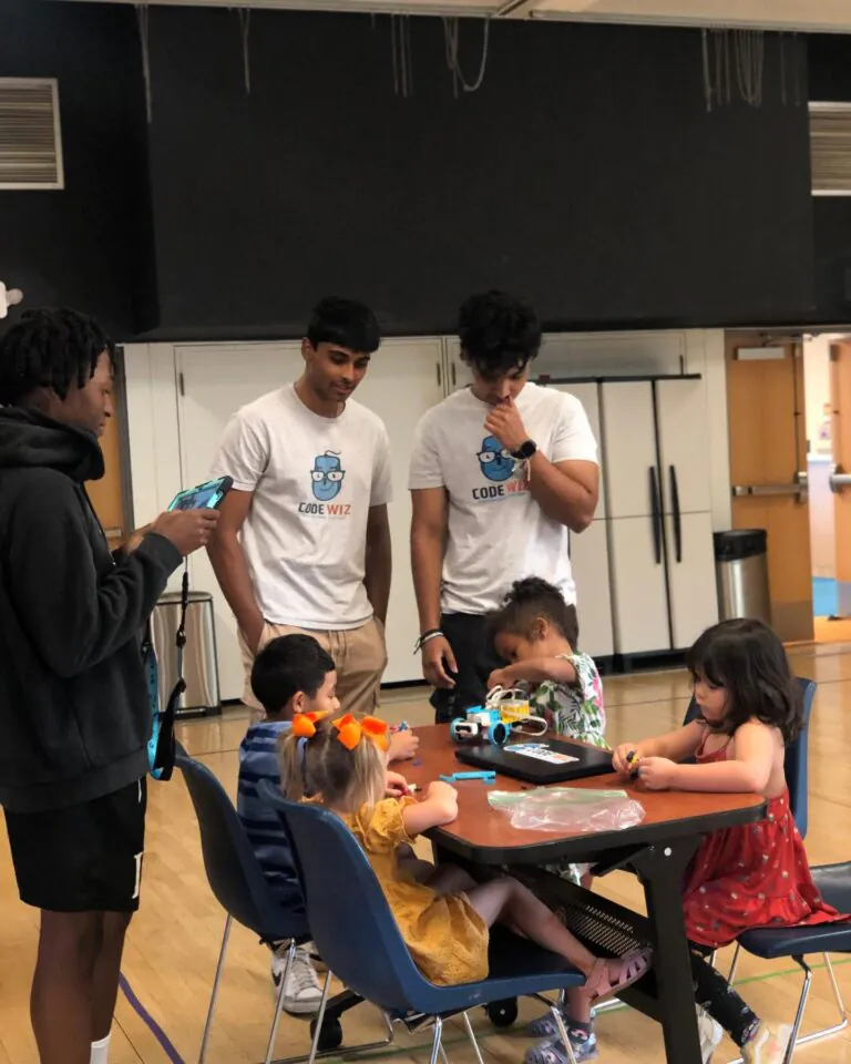 Young men in Code Wiz shirts teach children robotics at a table. TITLE: