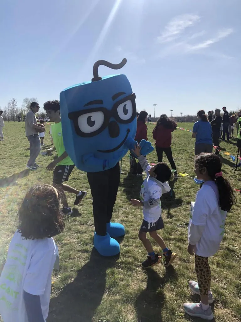 A friendly blue mascot with glasses interacts with a young child at an outdoor event on