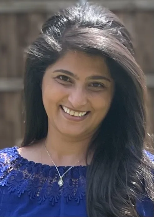 A smiling woman with dark hair wears a blue lace top and silver necklace against a blurred background.