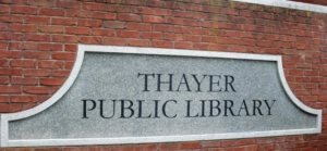 A gray granite sign displaying 'THAYER PUBLIC LIBRARY' is set into a red brick wall