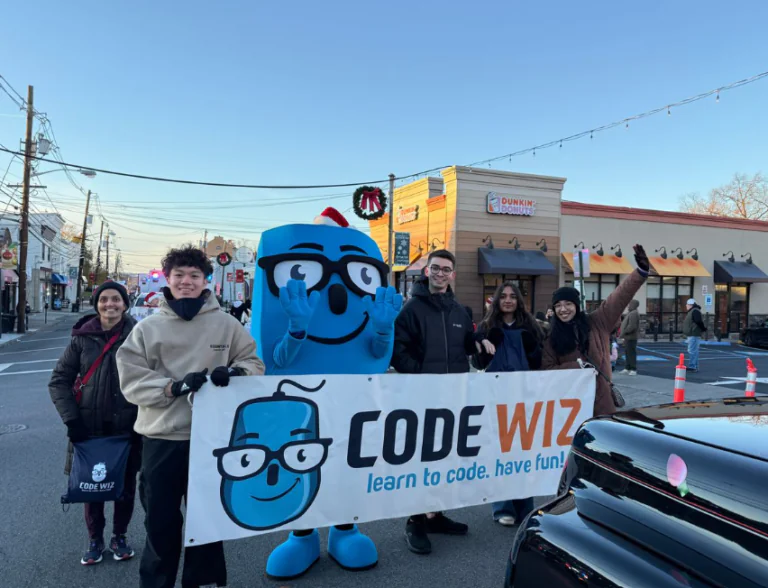 A Code Wiz mascot and five people hold a banner on a festive street with buildings in the background