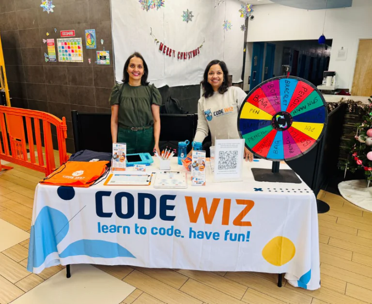 Smiling women promote Code Wiz coding classes at a holiday event with a colorful prize wheel.