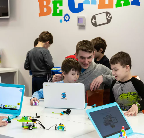 An adult guides two young boys using laptops and building robots in a tech classroom.