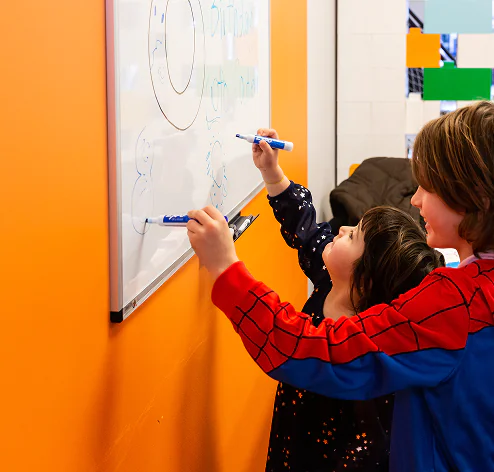 Two children, one in a Spider-Man hoodie, happily draw on a whiteboard with