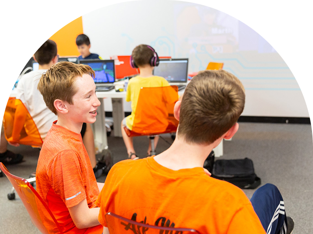 Smiling boys in orange shirts engage with laptops and each other in a modern learning environment
