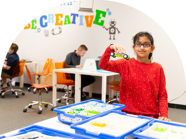 A smiling student holds a green block-built car in a creative classroom with other