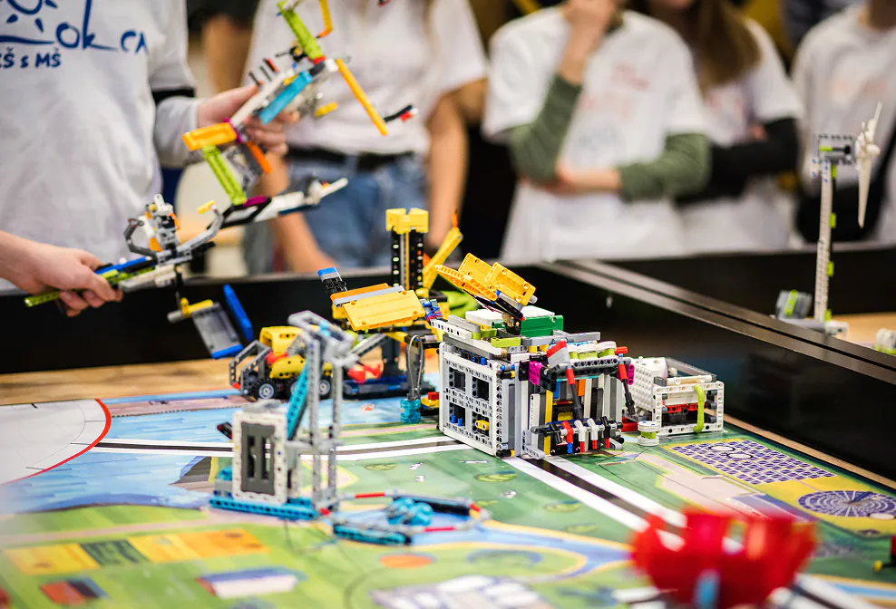 Students operate colorful robots on a patterned game mat during an engaging robotics competition. TITLE: