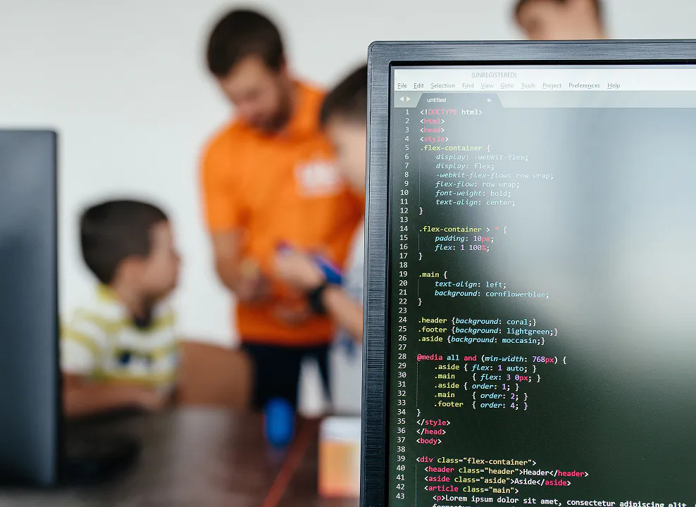 HTML and CSS code is displayed on a monitor with a blurred coding class in the background