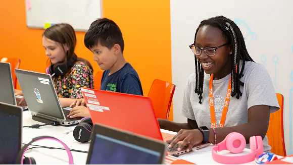 Diverse students of varying ages happily learn coding and technology on laptops in a bright classroom. TITLE